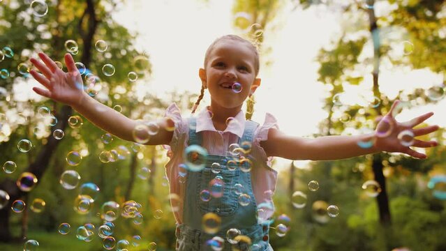 Cheerful little girl having fun while playing with soap bubbles flying in air, playful child catches bubbles in park outdoors, kid dream of happiness, happy childhood
