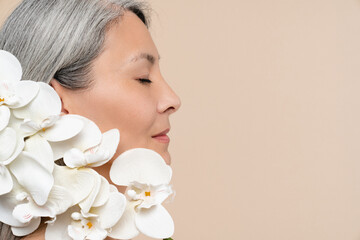 Closeup cropped shot flowers on face. Beauty concept. Caucasian pretty naked shirtless mature middle-aged woman with grey hair holding covering her face with orchid flower branch for aging skin care.