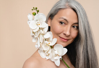 Beautification concept. Anti-age anti-wrinkle cosmetics beauty products for mature middle-aged naked shirtless woman covering her aging skin and face with orchid flower branch isolated in beige