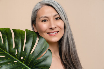 Beauty concept. Pretty caucasian mature middle-aged grey-haired woman with tropical leaf close to her face isolated in beige background. Ani-age cosmetics, rejuvenation aging skin