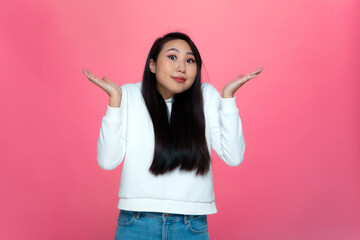 Confused positive puzzled young asian girl shrugged shoulders raised palms dont know answer on pink studio background