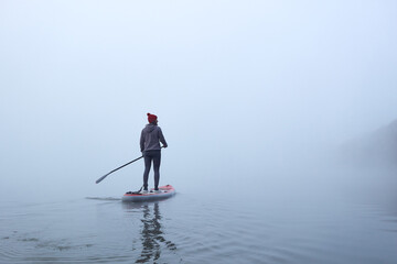 Alone woman rowing stand up paddle board (SUP) at misty Danube river at foggy autumn morning