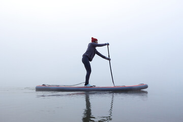 Alone woman rowing stand up paddle board (SUP) at misty Danube river at foggy autumn morning