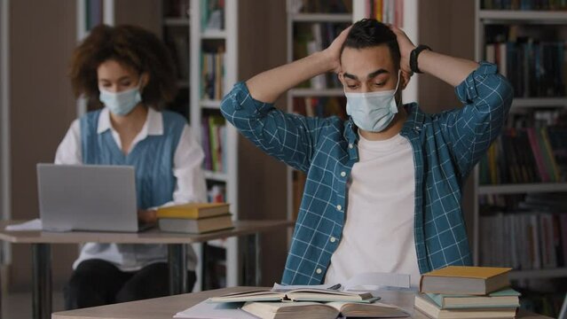 Tired overworked young arab male student in protective mask sits at desk in library does homework writes notes prepares for exam makes mistake grabs head with hands feels shock frustration failure