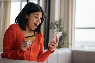 Overjoyed African American woman holding credit card using mobile phone shopping online at home....