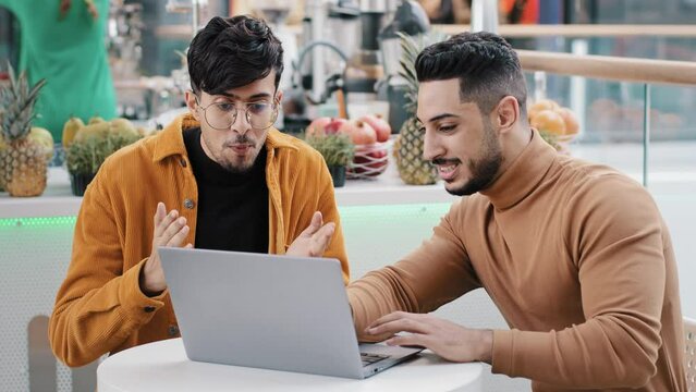 Young motivated arab guy demonstrates friend new application achievements on laptop indian man excitedly admiring work done applauding says wow showing thumbs up gesture approval supports colleague