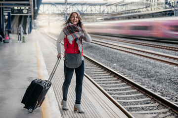 a smiling Caucasian woman in a vest and scarf waits for a train at a station while talking on the...
