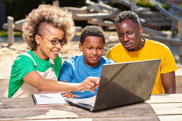 Portrait of happy afro parents and cute little boy using laptop together, watching photos or movie, browsing internet, choosing gifts outdoors
