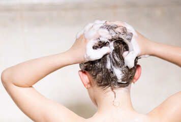 Little, beautiful girl, baby bathes sitting in a white bath with soap suds, shampoo and washes her head with hair. With your back to the camera
