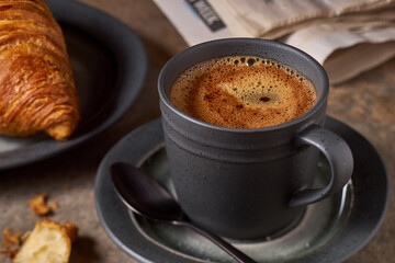 Black coffee with foam in dark cup and croissant on table