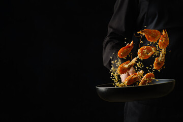A professional chef in a black uniform prepares king prawns in a frying pan on a black background....