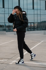 A young beautiful girl in a black short down jacket and sweatpants stands near a mirror wall. Long haired woman stands near a building and looks down.