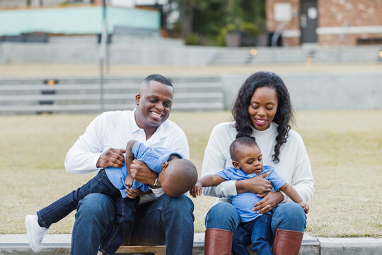 A beautiful African-American mom and dad sitting on steps outdoors and tickeling their two sons and everyone is laughing