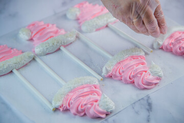 A pastry chef's hand in a transparent glove sprinkle coconut shavings on a pink meringue cap on a stick that lies on white parchment. High quality photo