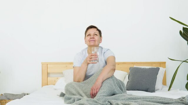 Smiling beautiful senior adult woman drinks water sitting in bed