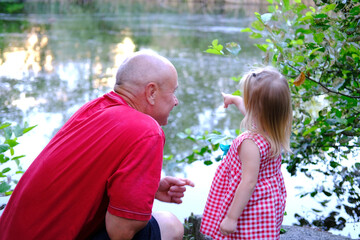 Naklejka premium small child, 2-year-old blonde girl in red dress, old man, grandfather and granddaughter by water, concept of childhood, family relationships of generations, walk in summer in nature