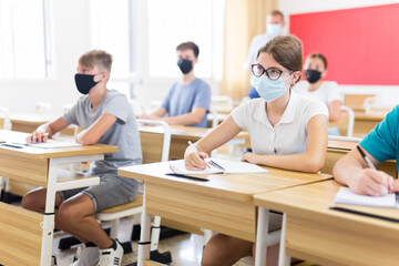 Teenagers in face masks sitting in class room. Male teacher explaining something to them.