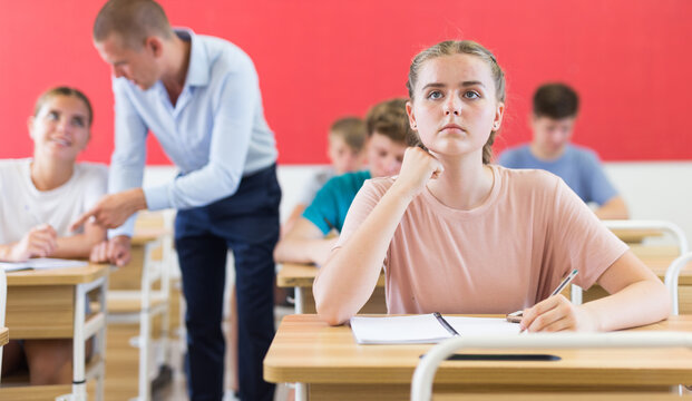 Diligent teenage girl studying in college with classmates, making notes of teacher lecture..