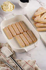 Step by step process of preparing italian dessert tiramisu out of ladyfinger biscuits, mascarpone cheese, espresso on white marble background