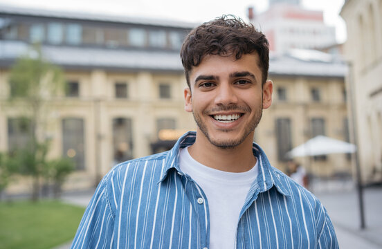 Portrait of smiling handsome Indian man wearing casual clothing standing on the street. Happy successful student looking at camera in university campus, education concept 