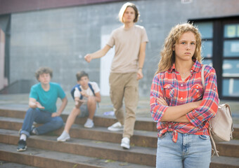 Sad young girl with backpack standing near school building. She's offended by her friends who trying to talk with her in background.