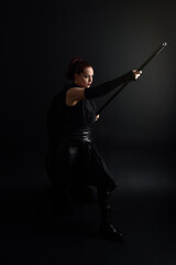 Full length portrait of pretty redhead female model wearing black futuristic scifi leather cloak costume. Standing pose on dark studio background with shadow rim  moody lighting.