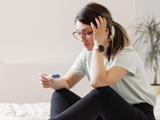 the woman found out about an unwanted pregnancy. A woman with a pregnancy test in her hands is sitting on the bed in her apartment - obrazy, fototapety, plakaty