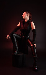 Full length portrait of pretty redhead female model wearing black futuristic scifi leather cloak costume. Sitting  pose on dark studio background with shadow rim  moody lighting.
