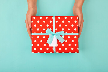 Hands give a red polka dot gift box with a card and a bow on isolated pastel blue-turquoise...