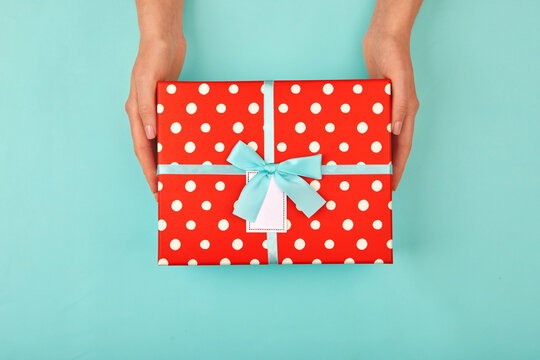Hands give a red polka dot gift box with a card and a bow on isolated pastel blue-turquoise background top view