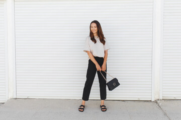 Young beautiful woman, wearing beige t-shirt, black pants, bag with chain and flat sandals walking outdoor near white roller door. Stylish trendy basic minimalistic casual outfit. Street fashion.