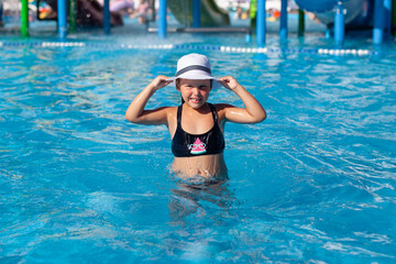 Obraz na płótnie Canvas Child is resting in water park. Little happy tanned girl in black swimsuit poses in pool and holds white straw hat decorated with black ribbon. 