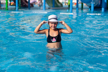 Child is resting in water park. Little happy tanned girl in black swimsuit poses in pool and holds white straw hat decorated with black ribbon. 