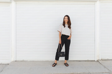 Young beautiful woman, wearing beige t-shirt, black pants, bag with chain and flat sandals walking outdoor near white roller door. Stylish trendy basic minimalistic casual outfit. Street fashion.