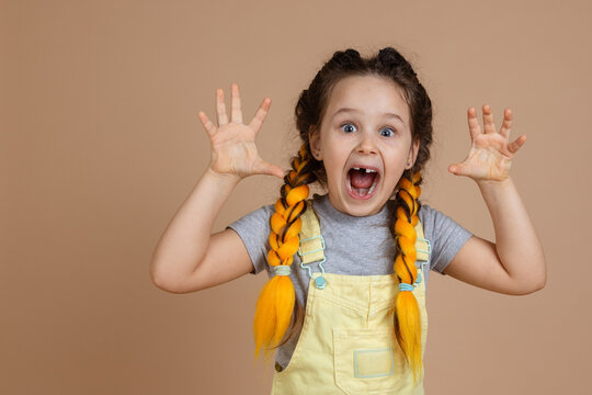 Portrait of indulging little girl having yellow kanekalon pigtails, frightening someone with hands and weird grimace wearing yellow jumpsuit and gray t-shirt on beige background.