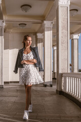 Obraz premium An attractive woman celebrates life in a summer white dress and denim jacket on the balcony of a summer resort. Joy and serenity