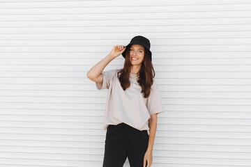 Young beautiful woman, wearing beige t-shirt, black pants, bucket hat and gold chain necklace, standing outdoor near white roller door. Stylish trendy basic minimalistic casual outfit. Street fashion.