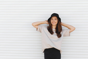 Young beautiful woman, wearing beige t-shirt, black pants, bucket hat and gold chain necklace, standing outdoor near white roller door. Stylish trendy basic minimalistic casual outfit. Street fashion.