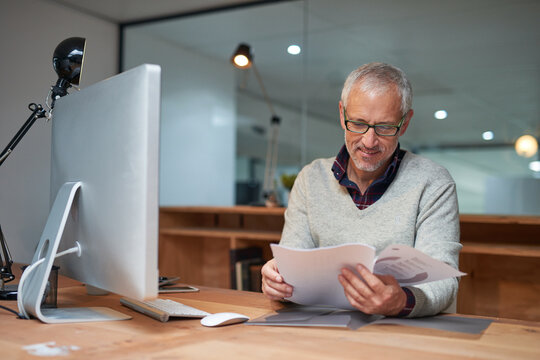 He always reads the fine print. Shot of a smiling mature businessman reading paperwork while sitting at his desk in an office.