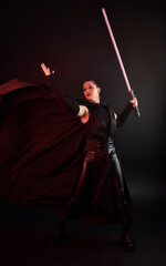 Full length portrait of pretty redhead female model wearing black futuristic scifi leather cloak costume. Standing pose  holding lightsaber on dark studio background with shadow moody lighting.