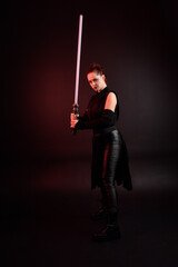 Full length portrait of pretty redhead female model wearing black futuristic scifi leather cloak costume. Standing pose  holding lightsaber on dark studio background with shadow moody lighting. - obrazy, fototapety, plakaty