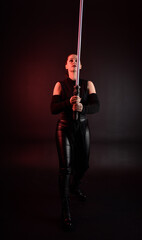 Full length portrait of pretty redhead female model wearing black futuristic scifi leather cloak costume. Standing pose  holding lightsaber on dark studio background with shadow moody lighting.