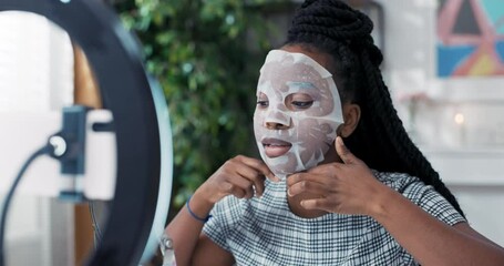 A beautiful young African-American woman is enjoying beauty treatments at home, applying a sheet mask to her face. She records a skin care tutorial for her social media channel.
