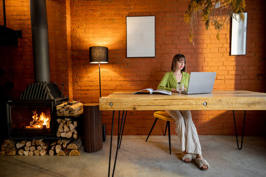 Woman works on laptop while sitting by the wooden table in cozy living room with a burning fireplace. Interior in loft style
