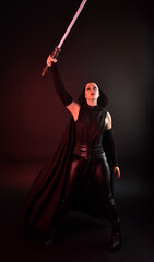 Full length portrait of pretty redhead female model wearing black futuristic scifi leather cloak costume. Standing pose  holding lightsaber on dark studio background with shadow moody lighting.