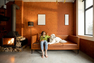 Young woman relax and listen to the music while sitting with her cute white dog on a couch at home....