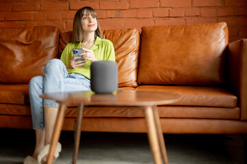 Obraz premium Young woman using phone while sitting relaxed on leather couch with smart speaker in front. Concept of smart home and leisure time with gadgets
