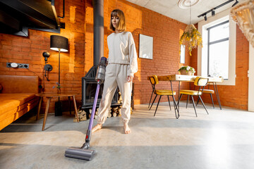 Woman cleaning concrete floor with cordless handheld vacuum cleaner in living room at home. Housewife doing housework - obrazy, fototapety, plakaty