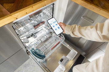 Woman holding phone with running smart home application, controlling dishwasher remotely. Smart...