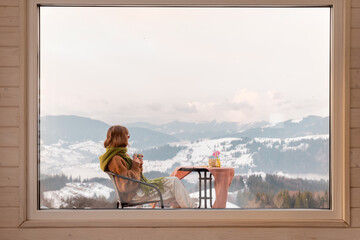 Woman sitting with a drink on terrace of tiny house in the mountains, view through the window from...